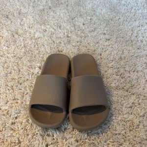 Beige slides med platt sula - Säljer ett par stilrena slides i beige färg med platt sula och öppen tå. De har en bred rem över foten och är tillverkade i syntetmaterial med lätt textur. Perfekta för chill dagar och enkel att dra på sig. Sulan har mönstrad undersida för extra grepp. Storlek 37-38 