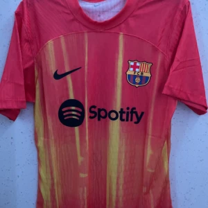 Barcelona Lamine Yamal #10 Nike tröja - Säljer en Barcelona fotbollströja med Lamine Yamal och nummer 10 på ryggen. Tröjan är röd med gula ränder, har korta ärmar och är tillverkad i ett lätt, ventilerande material. Nike-logga och Spotify sponsortryck på framsidan samt Barca-emblem på bröstet.