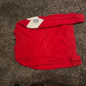 Röd långärmad tröja från Moncler - Snygg röd långärmad tröja från Moncler Gamme Bleu med vit patch och broderad logga på ena ärmen. Rund hals och ribbade muddar. Tillverkad i mjuk bomull, perfekt för dig som gillar stilrena detaljer och exklusiva märken. 