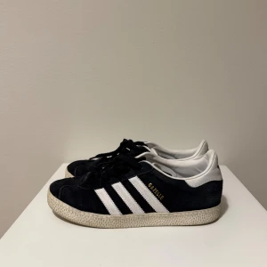 Adidas Gazelle sneakers mocka - Adidas Gazelle sneakers med klassiska vita ränder och vit häl. Snygg låg modell med rund tå och platt sula. Ovandel i mjuk mocka och snörning framtill. Perfekt för dig som gillar retrostil och streetwear.