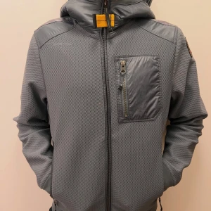 Grå vindjacka från Parajumpers - Snygg grå vindjacka från Parajumpers med huva och coolt badge på ärmen. Jackan har quiltade detaljer på axlar och huva, dragkedja framtill och en praktisk bröstficka med zip. Materialet är polyester och syntet, perfekt för blåsiga dagar.