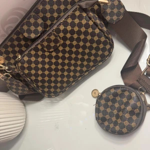 Brun axelväska  - Snygg brun axelväska med guldigt mönster och bred axelrem. Väskan har flera fack med dragkedja och en matchande rund myntficka som hänger på sidan. Materialet är syntet och detaljerna är i guld. Perfekt för dig som vill ha en trendig och praktisk väska.