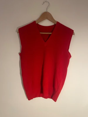 Röd stickad väst med v-ringning - Riktigt snygg röd stickad väst med v-ringad hals. Västen har en klassisk, enkel design utan mönster och ärmar. Perfekt att styla över en skjorta eller t-shirt för en cool look. Ribbad kant vid hals och lite sönder i vänstra arm är sista bilden