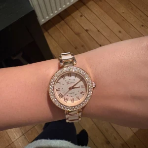 Glittrig guldig klocka  - Säljer en snygg klocka  med glittrande stenar runt urtavlan och guldiga detaljer. Urtavlan är rund med romerska siffror och armbandet har vita länkar i kombination med guld. 