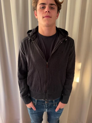 Svart cardigan Polo Ralph Lauren - Svart vindjacka från Polo Ralph Lauren med huva och dragkedja framtill. Jackan har diskret logga på bröstet, detaljer i syntetmaterial på axlar och huva samt ribbad nederkant. Perfekt för dig som vill ha en stilren och enkel jacka med sportig känsla.