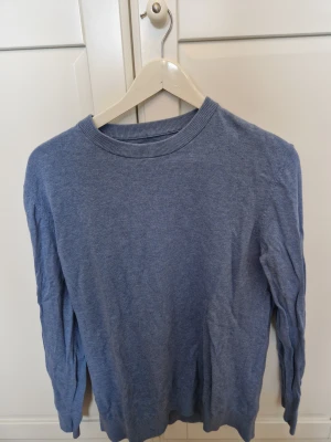 Blå stickad tröja med rund hals - En enkel och snygg blå stickad tröja med rund hals och långa ärmar. Perfekt för dig som gillar en clean look och vill ha något mjukt och bekvämt. Tröjan har ribbade muddar vid hals, ärmslut och nederkant. Passar till jeans eller chinos.