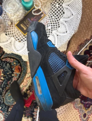 Nike Air Jordan 4 svart/blå sneakers - Säljer ett par Nike Air Jordan 4 sneakers i svart med blå detaljer. Skorna har meshpaneler, snörning och en synlig luftbubbla i sulan. Ikonisk Jumpman-logga på plösen och cool design med kontrasterande färger. Perfekt för dig som gillar streetwear och basketstil.