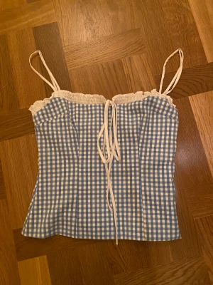 Blåvit rutig linnetopp med knyt - Supersöt populär linnetopp med blåvitt rutigt mönster och tunna vita axelband. Toppen har en söt volangkant upptill och knytning framtill för extra detalj. Perfekt för varma dagar och ger en fräsch och ungdomlig vibe. Materialet känns lätt och luftigt. Använts två gånger🩵