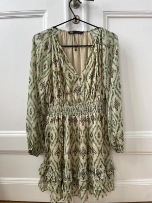 Grön mönstrad kortklänning från Zara - Säljer en superfin kortklänning från Zara med grönt och beige mönster, v-ringad hals och långa ärmar. Klänningen har smock i midjan och volanger nedtill för extra flow. Perfekt för dig som gillar boho-vibe och snygga detaljer.
