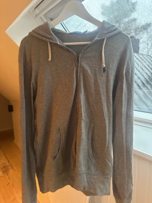 Grå zip hoodie från Polo Ralph Lauren - Snygg grå hoodie från Polo Ralph Lauren med dragkedja, huva och vita snören. Tröjan har långa ärmar, två fickor framtill och den klassiska lilla Polo-loggan broderad på bröstet. Knappt använd