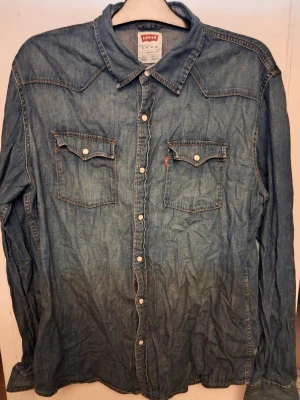 Blå jeansskjorta från Levi's XL - Snygg blå jeansskjorta från Levi's i storlek XL. Klassisk westernstil med två bröstfickor med knappar och coola detaljer på axlarna. Skjortan har långa ärmar och knappar framtill. Perfekt för dig som gillar avslappnad denim-look.