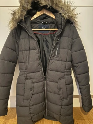 Mörkgrå pufferjacka med fuskpäls - Snygg mörkgrå pufferjacka från ONLY i storlek XS. Jackan har en fluffig huva med fuskpäls, quiltad design och praktiska fickor med knappar. Dragkedja framtill och en extra ficka med zip på bröstet. Perfekt för kalla dagar och riktigt cozy vibe.