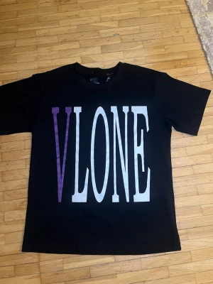 Svart VLONE t-shirt med lila tryck - Svart t-shirt från VLONE med stort tryck på bröstet i vitt och lila samt en stor lila V på ryggen. Klassisk rund hals och korta ärmar. Materialet är mjuk bomull som känns skön mot huden. Perfekt för dig som gillar streetwear och vill sticka ut.
