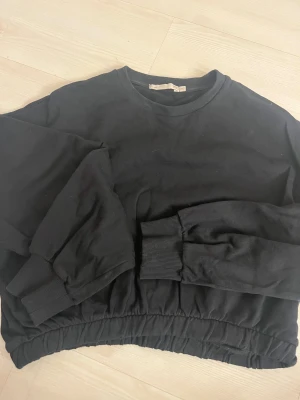 Svart croppad sweatshirt med resår - Svart croppad sweatshirt från NA-KD med rund hals och långa ärmar. Tröjan har breda muddar vid ärmslut och en resår i nederkant som ger en snygg, avslappnad siluett. Perfekt för en chill och trendig look.