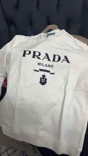 Prada Milano - Vit sweatshirt från Prada Milano med svart tryck på bröstet och rund hals. Tröjan har långa ärmar och klassisk passform. Materialet är mjuk bomull och den har en stilren look med tydlig logga framtill. Perfekt för dig som gillar exklusiva märken och enkel design.
