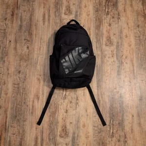 Svart ryggsäck från Nike - Svart ryggsäck från Nike med stort logotryck i silver på framsidan. Väskan har flera fack med dragkedja, justerbara och vadderade axelremmar samt ett praktiskt bärhandtag upptill. Materialet är slitstark polyester och designen är stilren och sportig.