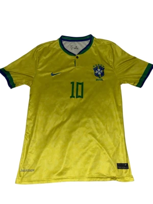 Brasilien Neymar Jr. 10 fotbollströja - Gul Brasilien fotbollströja från Nike med gröna detaljer och blå Nike-logga. Nummer 10 och Neymar Jr. tryckt på ryggen. CBF-emblem på bröstet, korta ärmar med grönt och blått kantband. Tillverkad i lätt, ventilerande material med Dri-Fit ADV-teknologi.