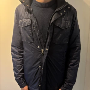 Mörkblå puffer/fieldjacka från J.Lindeberg - Snygg mörkblå pufferjacka från J.Lindeberg med två stora bröstfickor med lock, dragkedja och tryckknappar framtill. Jackan har lång ärm och patch med JL-logga på vänster arm. Yttermaterialet är polyester och fodret är mjukt och varmt. Funkar till både M och L! Priset går att diskutera!