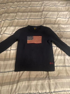 Mörkblå stickad tröja Polo Ralph Lauren - Snygg mörkblå stickad tröja från Polo Ralph Lauren med amerikansk flagga broderad på bröstet och RL-broderi i rött vid nederkanten. Tröjan har rund hals och långa ärmar med ribbade muddar. Perfekt för dig som gillar klassisk stil med en twist.
