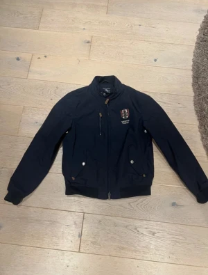 Mörkblå bomberjacka Morris Gents M - Snygg mörkblå bomberjacka från Morris Gents med broderad logga på bröstet och Union Jack-flagga på insidan. Jackan har dragkedja, ståkrage med knapp och två fickor med tryckknappar. Perfekt för dig som gillar klassisk brittisk stil.