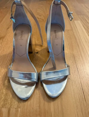Silvriga klackpumps från Bershka - Säljer ett par snygga silvriga pumps från Bershka i storlek 36. Skorna har en bred klack, öppen tå och rem runt vristen med spänne. Perfekt för dig som gillar metallic och vill sticka ut. Materialet är syntet med glansig finish.