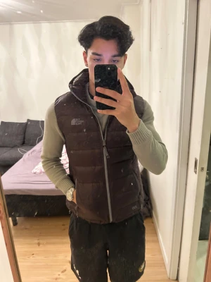 Mörkbrun dunväst från The North Face - Snygg mörkbrun dunväst från The North Face med broderad logga på bröstet. Västen har huva, två sidofickor med dragkedja och quiltad design. Perfekt för lager-på-lager och har en stilren look med 700-fill märkning på nederkanten.