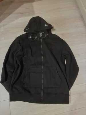 Svart hoodie med goggles  - Svart hoodie med ikoniska goggles i huvan och broderad logga på bröstet. Jackan har dragkedja framtill, två stora fickor och coola metalldetaljer på huvan. Perfekt för dig som vill sticka ut med streetwear-vibe.