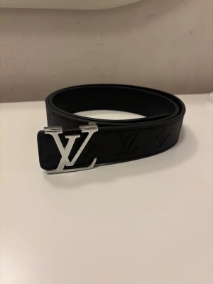 Svart Louis Vuitton skärp i skinn - Snyggt svart Louis Vuitton bälte i skinn med präglade LV-loggor och klassisk silverfärgad LV-spänne. Bältet är brett och har en stilren design som ger en lyxig vibe. Perfekt för dig som vill ha en statement-accessoar med exklusiv känsla.