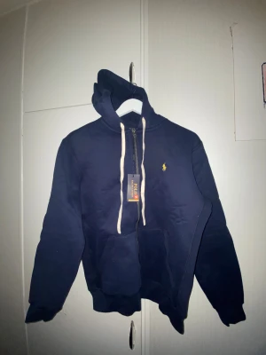 Marinblå hoodie från Polo Ralph Lauren - Snygg marinblå hoodie från Polo Ralph Lauren med gul broderad logga på bröstet. Tröjan har vit snörning i huvan, dragkedja framtill och en stor magficka. Perfekt för chill dagar och enkel att matcha med jeans eller joggers.