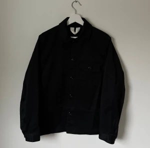 Svart jacka från ARKET, strl 46 - Svart jacka från ARKET i ekologisk bomullssatin. Klassisk krage, knappstängning framtill och en bröstficka med lock. Snygg, stilren och passar perfekt till lager-på-lager. Långärmad och med normal passform. 5H4