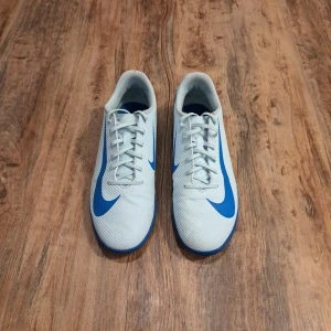 Nike fotbollsskor - Inomhus  - Säljer ett par snygga ljusblå Nike fotbollsskor med blå detaljer och logga. Skorna har snörning och en låg profil med platt sula, perfekt för inomhusspel. Materialet är syntet och designen är sportig med en modern touch. Stora Nike-tryck på hälen och sidan ger extra stilpoäng.