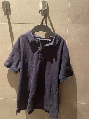 Marinblå pikétröja från GANT - Snygg marinblå pikétröja från GANT med klassisk krage och två knappar framtill. Tröjan har korta ärmar och en liten broderad logga på bröstet. Tillverkad i mjuk bomull som är skön mot huden. Perfekt för dig som gillar stilrena och tidlösa plagg.