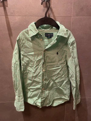 Grönvit randig skjorta från Ralph Lauren - Snygg skjorta från Ralph Lauren i grön och vit randigt mönster. Klassisk krage, långärmad och med knappar framtill. Ikonisk broderad logga på bröstet. Tillverkad i mjuk bomull som känns skön mot huden. Perfekt för dig som gillar stilrena och fräscha plagg.