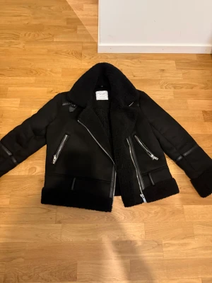 Svart liknande skinnjacka ifrån STRADIVARIUS  - Säljer en svart liknande skinnjacka från STRADIVARIUS med bred krage och mjukt foder. Jackan har silvriga dragkedjor, detaljer i skinnimitation och ribbade muddar. Perfekt för dig som vill ha en cool och varm look i höst och vinter.