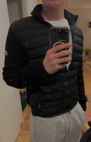 Svart Moncler pufferjacka med stickade ärmar - Svart pufferjacka från Moncler med stickade ärmar och hög krage. Jackan har quiltad framsida, två sidofickor med dragkedja och dubbla dragkedjor framtill. Moncler-logga på ärmen. Perfekt för dig som vill ha en clean och stilren look.