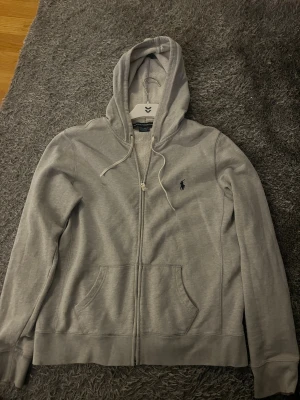 Grå hoodie från Polo Ralph Lauren - Snygg grå hoodie från Polo Ralph Lauren med dragkedja och huva med snörning. Tröjan har det klassiska Polo-logot broderat på bröstet och två praktiska fickor framtill. Perfekt för chill dagar och enkel att matcha med jeans eller joggers.