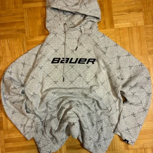 Grå hoodie från Bauer med hockeymönster - Grå hoodie från Bauer med svart logga på bröstet och all-over print av korsade hockeyklubbor. Tröjan har huva med snörning och är långärmad. Mjuk insida och klassisk loose passform, perfekt för chill eller sport. Materialet är bomull och polyester.