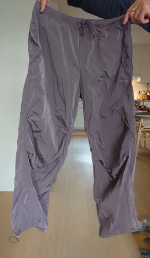 Lila/grå träningsbyxor från Freddy med snörning - Säljer ett par grå täckbyxor från Freddy med elastisk midja och snörning. Byxorna har en loose passform och är tillverkade i syntetmaterial som ger en lätt och skön känsla. Diskret Freddy-logga broderad på sidan och justerbara benslut med dragsko.