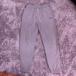 Grå mjukisbyxor från Celine M - Grå mjukisbyxor från Celine med elastisk midja och vit snörning. Byxorna har en clean look med Celine-logga på vänster höft och muddar vid bensluten. Perfekta för chill dagar och streetwear vibes. Nästan nyskick