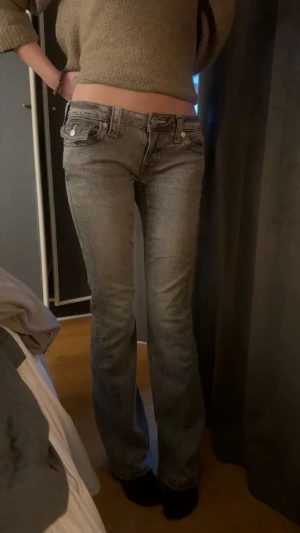 True Religion Joey Low Rise Flare Jeans - Säljer ett par gråa True Religion Joey jeans med låg midja och flare-ben. Klassiska detaljer med stora bakfickor, vita kontrastsömmar och silverfärgade knappar. Jeansen är i tvättad grå denim och har en snygg, avslappnad passform.