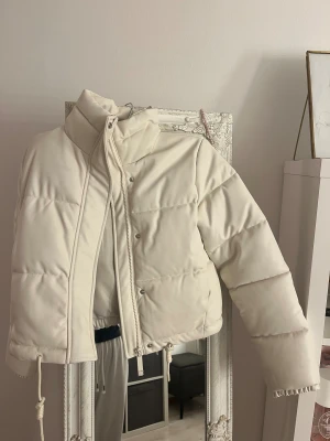 Vit pufferjacka med snörning - Säljer en snygg vit pufferjacka med hög krage och långa ärmar. Jackan har dragkedja och knappar framtill samt snörning i nederkant för att justera passformen. Materialet är mjukt och känns som syntet, perfekt för kalla dagar.