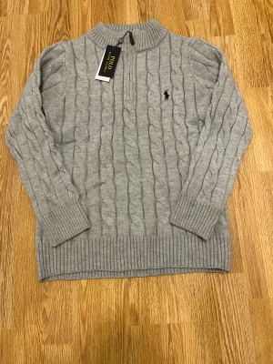 Grå stickad tröja Polo Ralph Lauren - Snygg grå stickad tröja från Polo Ralph Lauren med klassiskt kabelmönster och halv dragkedja vid halsen. Tröjan har ribbade muddar och det ikoniska broderade Polo-logot på bröstet. Perfekt för dig som gillar stilren och tidlös design.