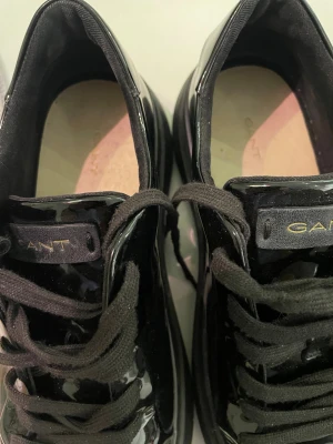 Svarta sneakers från GANT - Snygga svarta sneakers från GANT med glansig finish och klassisk snörning. Skorna har rund tå och platt sula, vilket ger en clean och modern look. Perfekta för dig som gillar stilrena och trendiga skor med lite extra attityd.