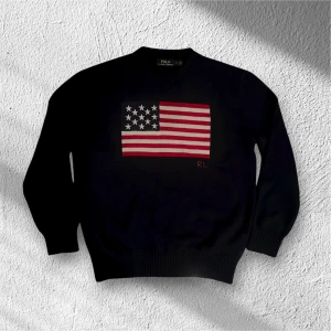 Mörkblå stickad tröja Polo Ralph Lauren - Mörkblå stickad tröja från Polo Ralph Lauren med amerikansk flagga broderad på bröstet och RL-detalj i rött. Rund hals och ribbade muddar vid ärmslut och nederkant. Perfekt för dig som gillar klassisk och cool stil.