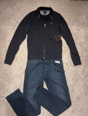 Mörk blå ziptröja från GANT - Mörk blå ziptröja från GANT med stilren design och diskret logga på bröstet. Tröjan har ståkrage, två fickor fram och silverfärgad dragkedja. Ribbade muddar vid ärmslut och nederkant. Perfekt för dig som gillar klassisk och sportig stil.