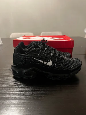 Nike Air Max Plus Tn Svarta sneakers - Nike Air Max Plus Tn sneakers i svart med vita detaljer och röd air-enhet. Ovandel i mesh och syntet, med tydliga Tn Air-loggor på häl och sula. Skorna har snörning, platt sula och en sportig, chunky design som sticker ut. Perfekt för dig som gillar streetwear och vill ha något unikt.