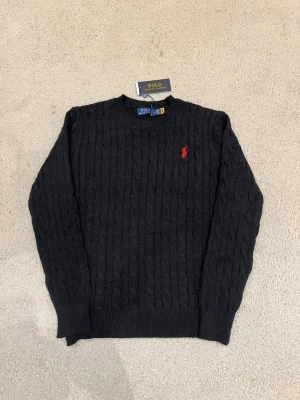 Ralph Lauren Sweatshirt  - Svart stickad tröja från Polo Ralph Lauren med klassiskt kabelmönster och röd broderad logga på bröstet. Rund halsringning och långa ärmar. Perfekt för dig som gillar stilrena och tidlösa plagg med en touch av lyx.
