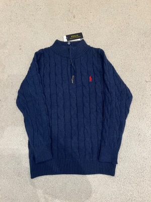 Ralph Lauren Zip Hoodie  - Snygg marinblå stickad tröja från Polo Ralph Lauren med klassiskt kabelstickat mönster och röd logga på bröstet. Tröjan har ribbad krage, muddar och half zip-dragkedja framtill. Perfekt för dig som gillar stilrena och tidlösa plagg.