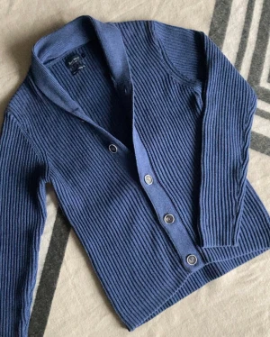 Ribbad kofta East West - Snygg blå kofta från East West med ribbad struktur och klassisk krage. Koftan har långa ärmar och stängs med  knappar framtill. Perfekt för dig som gillar stilrena och bekväma plagg med lite edge.