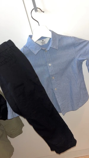Blå skjorta från H&M i bomull + svarta chinos  - Snygg blå skjorta från H&M i mjuk bomull. Klassisk krage, långa ärmar och knäppning framtill med vita knappar. Perfekt för en clean och stilren look. Svart Chinos byxor med knapp och resårband. 